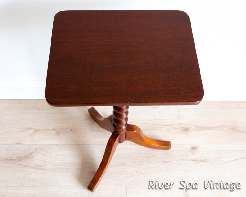 May include: A dark brown wooden side table with a square top and a twisted, turned, wooden pedestal base. The table is on a light wood floor.