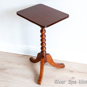 May include: A dark brown wooden side table with a square top and a turned, beaded pedestal base. The table has three legs that curve outward.