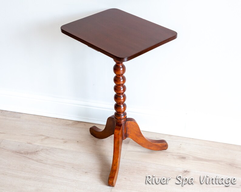 May include: A dark brown wooden side table with a square top and a turned, beaded pedestal base. The table has three legs that curve outward.