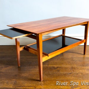 Puede incluir: Una mesa de centro de estilo moderno de mediados de siglo con un estante extraíble. La mesa está hecha de madera con un acabado marrón oscuro y tiene un estante de vidrio negro. La mesa está sobre un suelo de madera.