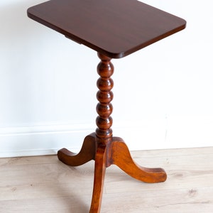May include: A dark brown wooden side table with a square top and a turned, beaded leg. The table has three curved legs and stands on a light wood floor.