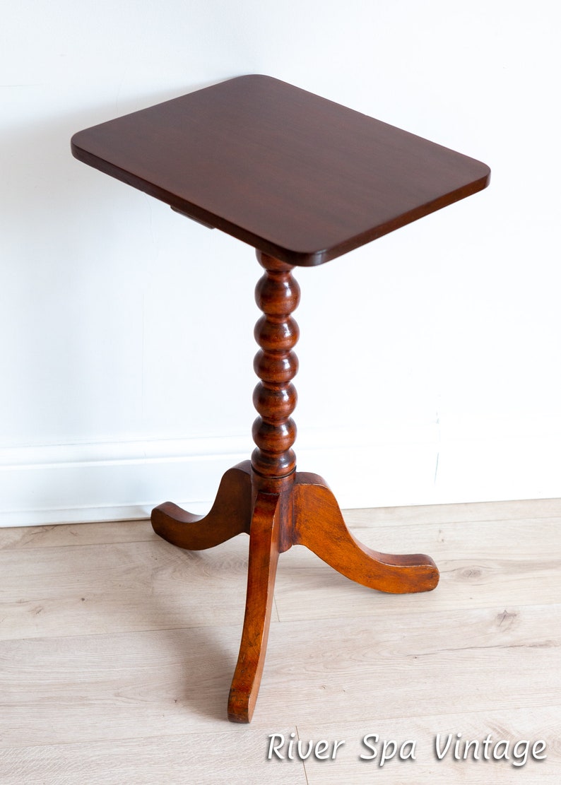 May include: A dark brown wooden side table with a square top and a turned, beaded leg. The table has three curved legs and stands on a light wood floor.