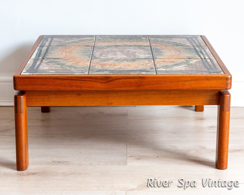May include: A mid-century modern coffee table with a tiled top featuring a geometric pattern in shades of brown, beige, and blue. The table has a teak wood frame and four legs.