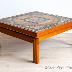May include: A square teak wood coffee table with a tiled top. The tiles are a mix of brown, green, and yellow with a circular pattern. The table has a simple design with four legs.
