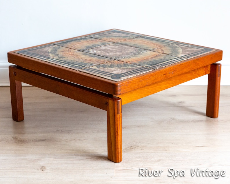 May include: A square teak wood coffee table with a tiled top. The tiles are a mix of brown, green, and yellow with a circular pattern. The table has a simple design with four legs.