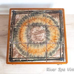 May include: A square, wooden coffee table with a tiled top. The tiles are brown, black, and orange with a swirling pattern. The table is in good condition and has a vintage look.