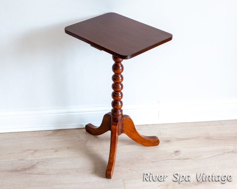 May include: A dark brown wooden side table with a square top and a turned, beaded leg. The table has a three-legged base with curved legs.