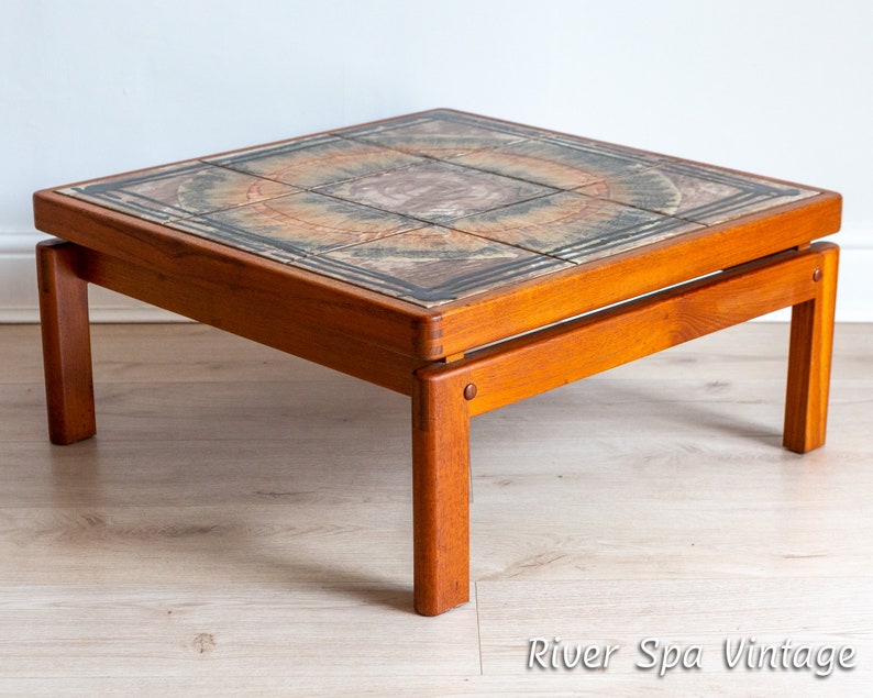 May include: A square, mid-century modern coffee table with a tiled top. The tiles are a mix of brown, orange, and green with a geometric pattern. The table has a solid wood frame and legs.