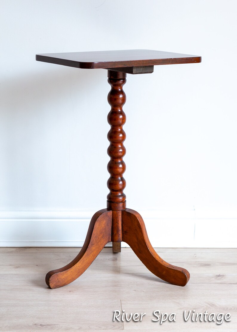 May include: A dark brown wooden side table with a square top and a turned, fluted pedestal base. The base has three curved legs.