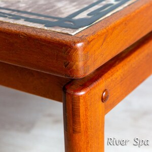 May include: Close-up of a wooden table with a tiled top. The table has a dark brown finish and the tile is a light brown with black accents. The table is made of solid wood and has a simple design.