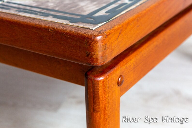 May include: Close-up of a wooden table with a tiled top. The table has a dark brown finish and the tile is a light brown with black accents. The table is made of solid wood and has a simple design.