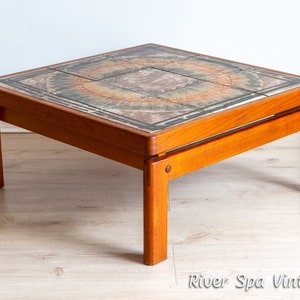 May include: A square teak wood coffee table with a tiled top featuring a circular pattern in shades of brown, green, and orange. The table has a simple, mid-century modern design.