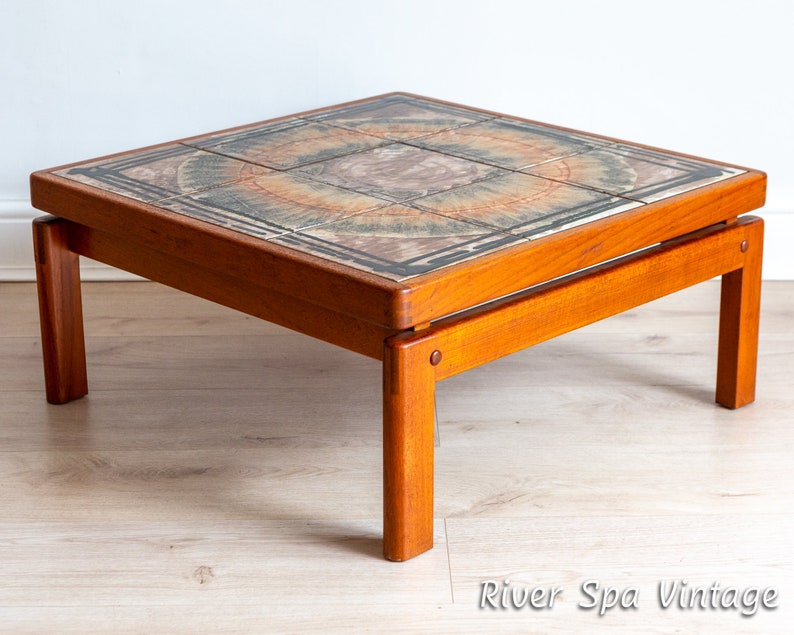 May include: A square teak wood coffee table with a tiled top featuring a circular pattern in shades of brown, green, and orange. The table has a simple, mid-century modern design.