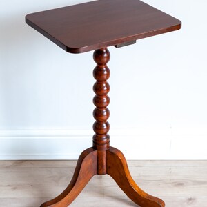 May include: A dark brown wooden side table with a round, beaded pedestal and a three-legged base. The table top is rectangular and has a smooth, polished finish.