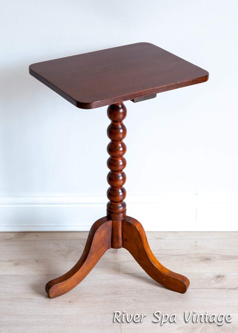 May include: A dark brown wooden side table with a round, beaded pedestal and a three-legged base. The table top is rectangular and has a smooth, polished finish.