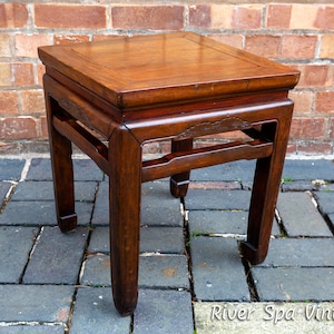 Op de afbeelding: Een donkerbruine houten bijzettafel met een vierkant blad en ingewikkelde houtsnijwerk. De tafel heeft vier poten met een uniek gebogen ontwerp. De tafel staat op een terras van bakstenen.