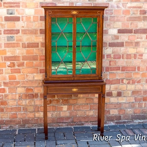 Puede incluir: Una vitrina de madera vintage con puertas de vidrio y un interior verde. La vitrina tiene un pequeño cajón y se apoya en cuatro patas. La vitrina está delante de una pared de ladrillo.