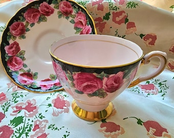 May include: A pink teacup and saucer with a black and gold rim and a floral design featuring pink roses. The teacup and saucer are on a white background with a floral pattern.