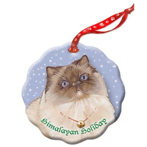 May include: A round ceramic Christmas ornament featuring a Himalayan cat with blue eyes. The ornament has a blue background with white snowflakes and the text "Himalayan Holiday" in green.