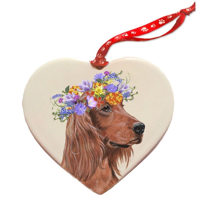 Op de afbeelding: Een keramische hartvormige ornament met een rode Ierse Setter die een kleurrijke bloemenkrans draagt. De ornament heeft een rood lint met witte poot afdrukken om op te hangen.