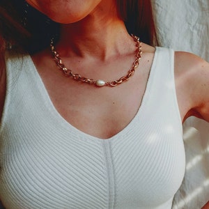 May include: A gold chain necklace with a large pearl pendant. The necklace is worn by a person wearing a white tank top.