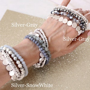 May include: A hand wearing three silver and gray beaded bracelets with silver disc charms. The bracelets are labeled "Silver-Gray", "Silver-GrayWhite", and "Silver-SnowWhite".