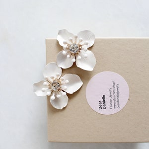 May include: A pair of white flower-shaped earrings with a rhinestone center. The earrings are packaged in a brown box with a pink circle sticker that says "Dear Danielle Fashion Jewelry www.etsy.com/shop/deardaniellejewelry"