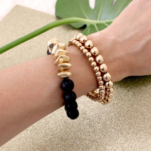 May include: Three gold beaded bracelets stacked on a wrist. The top bracelet features a gold geometric charm. The bottom bracelet is made of black lava beads.
