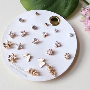May include: A white card with a variety of gold and pearl earrings. The earrings include butterflies, flowers, leaves, and simple pearl designs. The card has the text "Lead Compliant Designed in USA".
