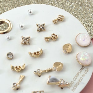 May include: A set of 14 gold-tone earrings with various designs, including keys, hearts, stars, and pearls. The earrings are displayed on a white card with the text "Lead Compliant Designed in USA".