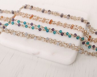 May include: Close-up of several strands of beaded jewelry. The beads are in various colors, including teal, purple, and cream, strung on gold-colored chains. The jewelry is displayed on a white surface.