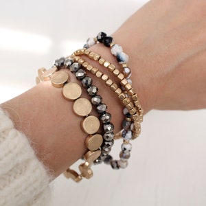 May include: A set of five bracelets featuring different styles and colors. The bracelets include gold beads, black and white agate beads, and silver-toned faceted beads. The bracelets are stacked on a person's wrist.