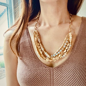 May include: A multi-layered gold necklace with white and beige beads. The necklace is made of metal and has a delicate, feminine design.