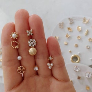 May include: A set of gold-toned earrings with various designs, including a star, a flower, a circle, and a pearl. The earrings are displayed on a white surface with a variety of other earrings.