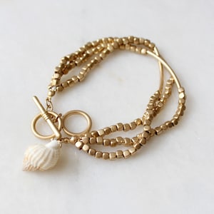 May include: A gold-toned bracelet with a seashell charm. The bracelet is made of multiple strands of small, square beads.