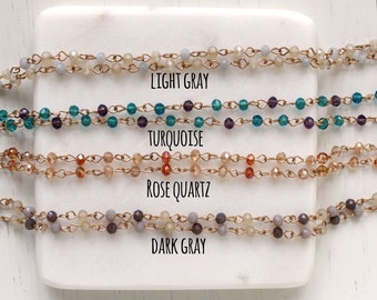 May include: Four strands of beaded necklaces with gold chain links. The beads are in four colors: light gray, turquoise, rose quartz, and dark gray. The necklaces are labeled with their corresponding colors.