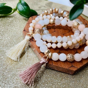 May include: Three layered bracelets with white and cream colored beads and gold accents. The bracelets have tassels on the ends, one white and one pink.
