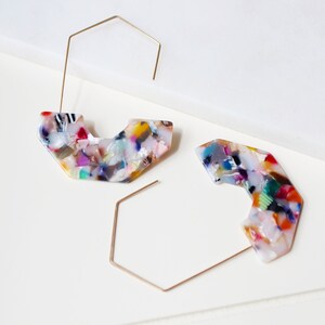 May include: A pair of gold hoop earrings with colorful, abstract, geometric, resin, statement pieces. The resin is white with multi-colored flecks.