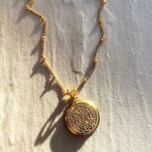 May include: A gold necklace with a round pendant featuring intricate details and a circular design. The pendant has a textured surface and appears to be made of metal.