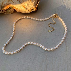 Peut inclure: Un collier délicat de perles avec un prolongement de chaîne en or orné d'un petit charme soleil. Le collier est composé de perles blanches et est posé sur une surface beige clair.