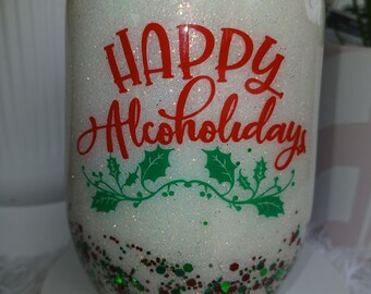 以下が含まれることがあります： 「HAPPY Alcoholidays」の赤い文字とヒイラギの葉のアクセントが入った白いグリッタートゥンブラー。タンブラーの底には赤と緑のグリッターアクセントがあります。タンブラーには黒い蓋が付いています。