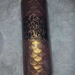 Puede incluir: Un vaso cilíndrico con un diseño de escamas de sirena en tonos dorados y morados. El vaso tiene el texto "Be a mermaid and make waves" en escritura negra. También es visible una silueta de cola de sirena.