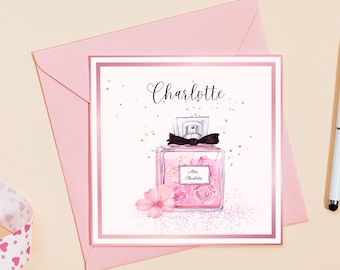 Puede incluir: Una tarjeta de felicitación rosa y blanca con un borde dorado. La tarjeta presenta una ilustración de acuarela de un frasco de perfume rosa con un lazo negro y flores. El texto "Charlotte" está escrito en cursiva negra en la parte superior de la tarjeta.
