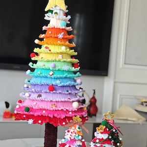 May include: A colorful, handmade Christmas tree with rainbow-colored tiers, topped with a star and Santa Claus figure. Two smaller decorative trees are also present, one with a gold star and the other with a green base. The trees are on a white table.