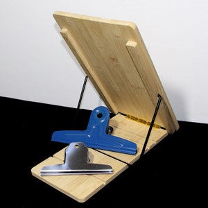 May include: A bamboo cookbook stand with a hinged design and black metal support. A blue and a silver metal binder clip rest on the stand. The stand is made of light-coloured bamboo.
