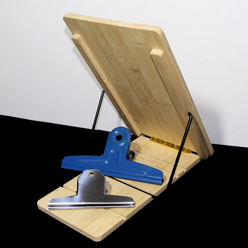 May include: A bamboo cookbook stand with a hinged design and black metal support. A blue and a silver metal binder clip rest on the stand. The stand is made of light-coloured bamboo.