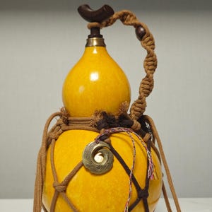 May include: A yellow gourd bottle with a capacity of about 550ml-600ml, labelled #51. It features a dark brown wooden stopper, a brass neck ring, and a decorative brass medallion. The gourd is encased in a brown macrame cord net with a handle.