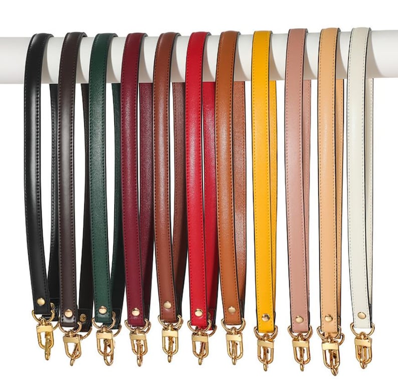 May include: A collection of leather bag straps in various colors, including black, brown, green, red, and yellow. Each strap is attached to a gold-colored metal clasp. The straps are hanging from a white bar.