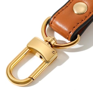 May include: Close-up of a gold-colored metal clasp with a brown leather strap. The clasp is a rectangular shape with a curved hook. The leather strap is secured with a gold-colored metal rivet.