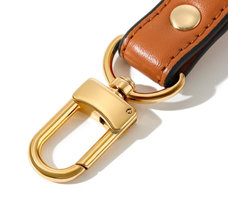 May include: Close-up of a gold-colored metal clasp with a brown leather strap. The clasp is a rectangular shape with a curved hook. The leather strap is secured with a gold-colored metal rivet.